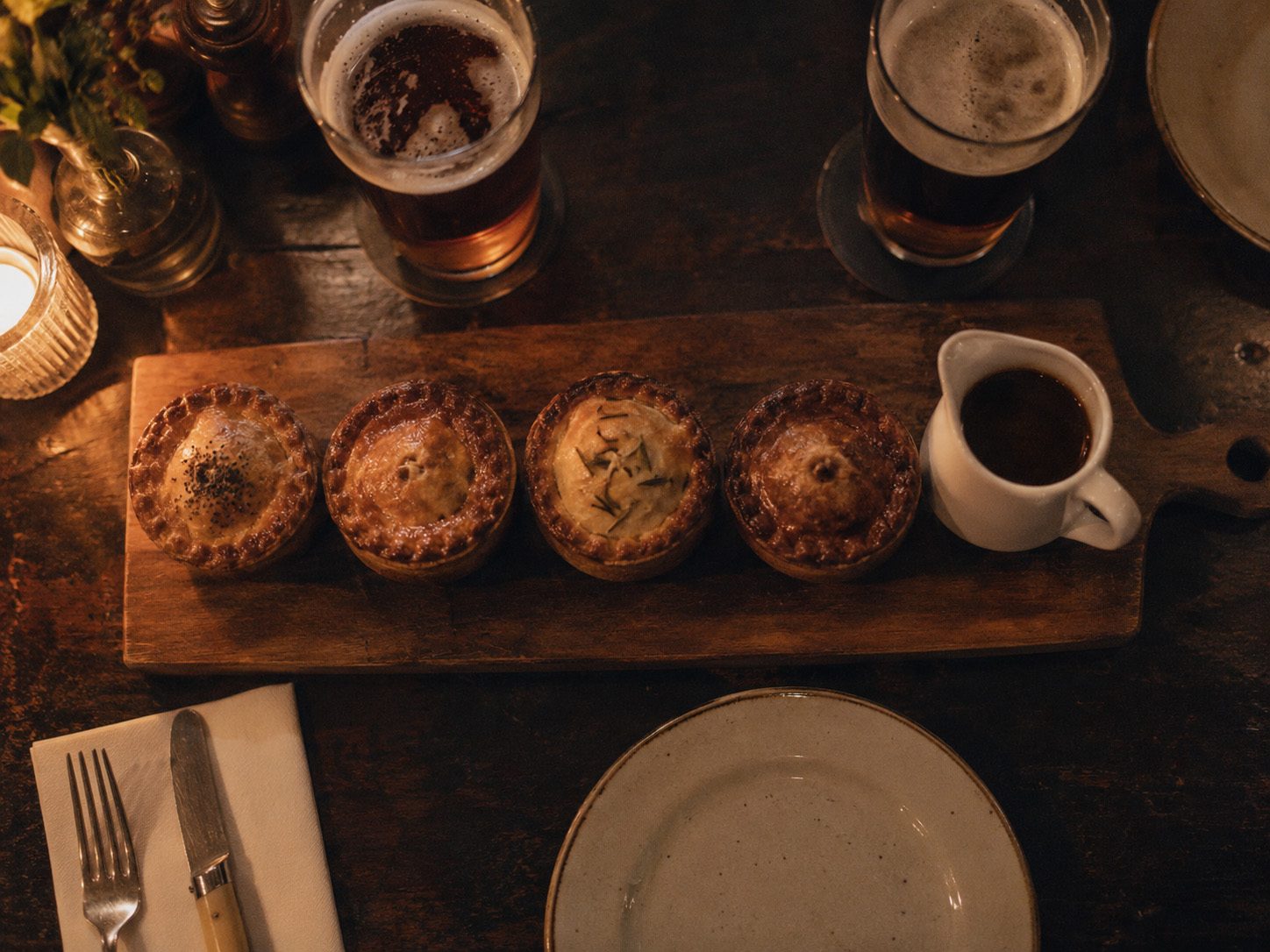 Concept image, generated: four small pies on a board with a jug of gravy and two pints, seen from above